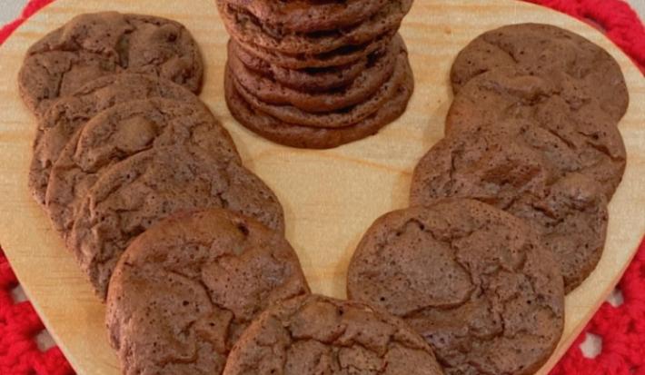 Fotografia de uma tábua de madeira em formato de coração sobre um pano vermelho. Sobre a tábua tem uma torre de uma receita de cookie brownie, ao lado de mais unidades do cookie completando o espaço da tábua.