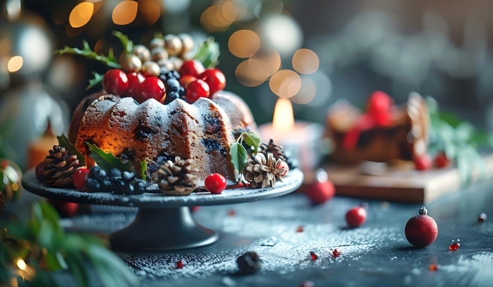 Fotografia de um bolo bundt decorado, com açúcar de confeiteiro e enfeites festivos como cerejas vermelhas e pinhas.
