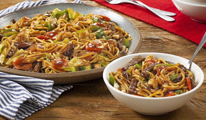 Fotografia em tons de marrom e vermelho de uma bancada de madeira, sobre ele um paninho azul e branco com um prato redondo com o yakissoba. A frente um recipiente redondo branco com yakissoba. Ao fundo um paninho vermelho.