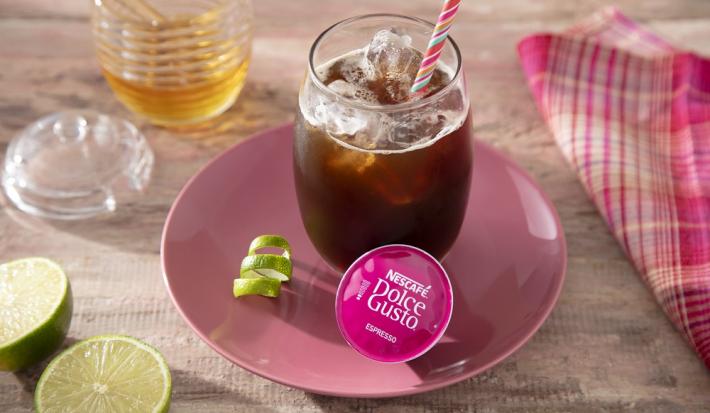 Foto em tons de for de rosa da receita de espresso gelado com limão e mel servida em um copo largo de vidro com bastante gelo em cima de um prato rosa de porcelana com uma raspa de limão e uma cápsula de dolce gusto espresso sobre uma mesa de madeira
