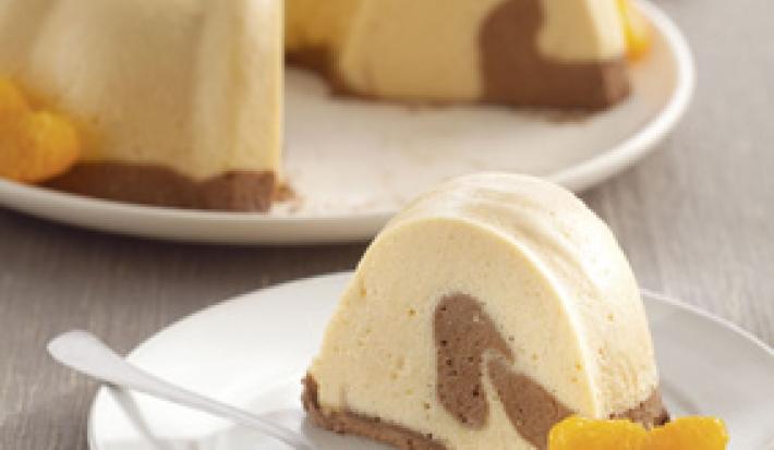 Fotografia em tons de laranja em uma bancada de madeira cinza, um prato branco redondo com a mousse de tangerina e chocolate em cima dele. Ao lado, um prato redondo branco de sobremesa com uma fatia da mousse.