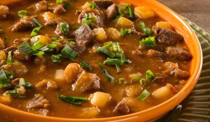 Foto aproximada de um prato marrom com a receita de carne de panela com mandioca dentro, em um caldo grosso e marrom, com pedaços de carne, mandioca e cheiro-verde picado