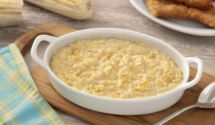 Foto da receita de Creme de Milho Cremoso. Observa-se um refratário branco com o creme de milho, do lado esquerdo superior espigas e do lado direito superior filés de frango empanados. O refratário do creme está sobre uma tábua de madeira.