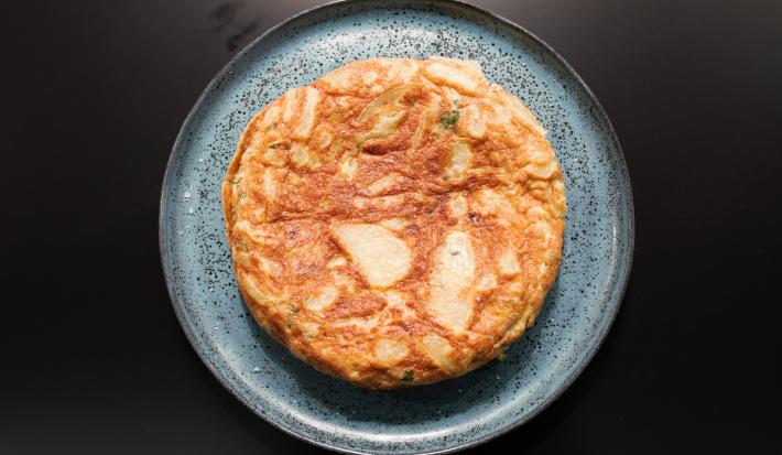 Fotografia em tons de preto com um prato azul ao centro. Dentro do prato existe uma receita de Tortilha Espanhola, uma receita feita com ovos e batatas cortadas de forma fina.
