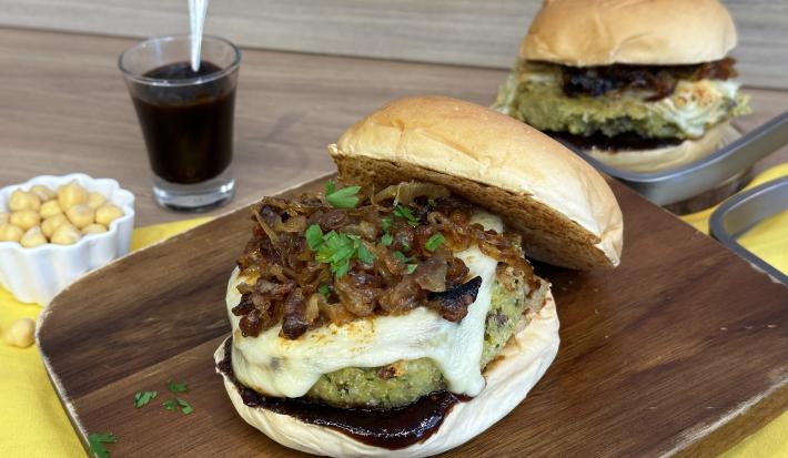 Fotografia de um hambúrguer completo com pão, cebola caramelizada, hambúrguer vegetal de cor esverdeada e queijo derretido. Ao fundo existe outro hambúrguer igual e ao lado um pequeno copo com molho de cor escura.
