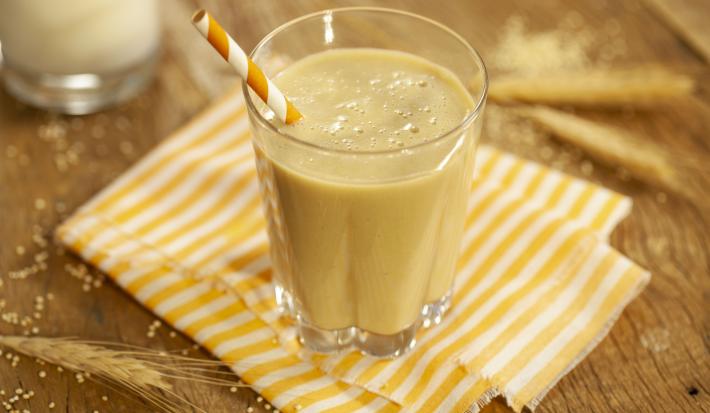 Fotografia em tons de marrom e amarelo de uma bancada de madeira com paninho listrado amarelo e branco, sobre ele um copo de vidro com a vitamina deliciosa e um canudo listrado amarelo e branco. Ao fundo ramos de trigo.