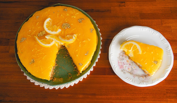 Fotografia mostra cheesecake de limão siciliano visto de cima, com cobertura amarelo vibrante e fatias de limão. Uma fatia servida ao lado em prato branco decorado.
