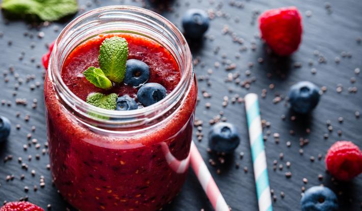 Fotografia de um copo de vidro decorativo sobre uma mesa preta, com frutas vermelhas e sementes de linhaça espalhadas ao lado de dois canudos. Dentro do copo tem um smoothie de frutas vermelhas com algumas blueberries e folhas de hortelã decorando.