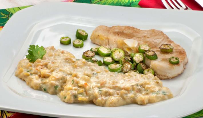 Fotografia em tons de verde em uma mesa com uma toalha verde florida, um prato branco grande com o peixe servido com quiabo e com purê de banana da terra.