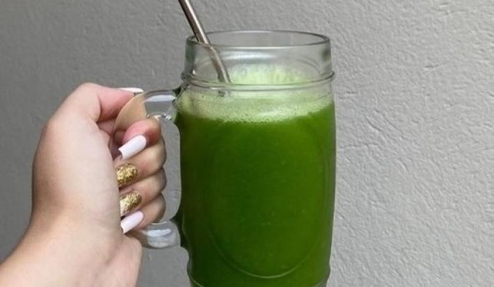 Fotografia de uma mão feminina segurando uma caneca de vidro grande com suco detox, de cor verde, com canudo de alumínio.