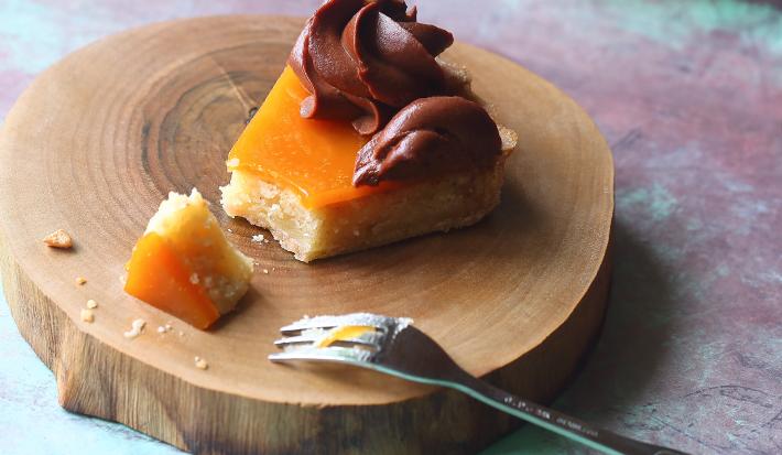 Fotografia de uma fatia de torta com damasco e mousse de chocolate por cima decorado por um bico de confeitar sobre uma tábua redonda de madeira e um garfo apoiado sobre. Ao fundo, a torta inteira sobre outra tábua de madeira redonda.
