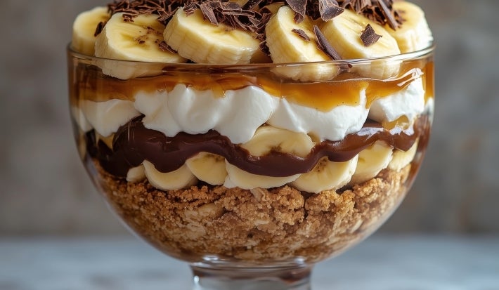 Em cima de uma mesa, refratário com biscoitos crocantes, doce de leite, bananas em rodelas, decorado com chantilly, bananas em rodelas e raspas de chocolate.
