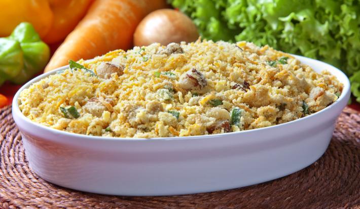 Fotografia em tons de amarelo em uma bancada de madeira de cor marrom. Ao centro, uma travessa branca contendo a farofa e ao fundo, alguns legumes e verduras espalhados.