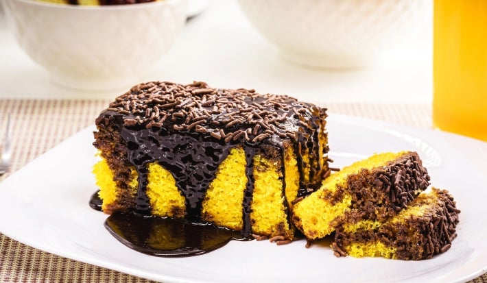 Pedaço de bolo de cenoura com cobertura de chocolate e granulado visto de outro ângulo. A calda escorre pelas laterais e se acumula no prato, criando um visual apetitoso. As fatias ao lado ainda aparecem, com tigelas brancas e um copo de suco.