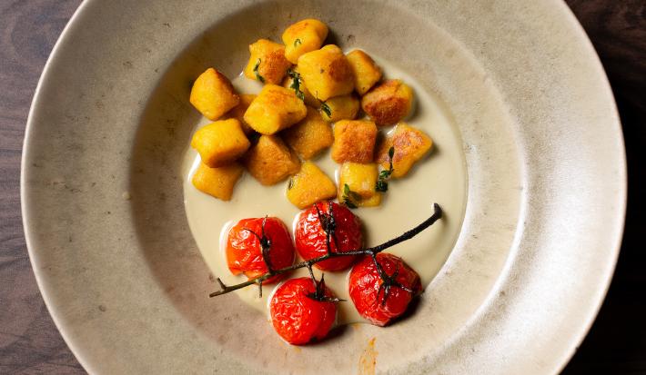 Fotografia em tons de branco com um prato branco ao centro. Dentro do prato existe uma receita de nhoque de abóbora acompanhada de tomates confitados