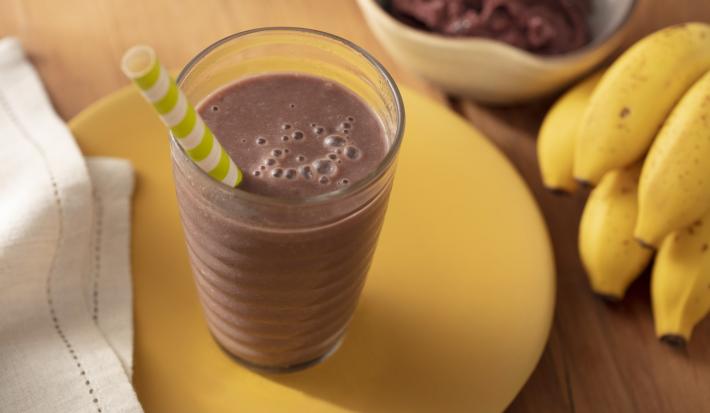 Fotografia em tons de amarelo e marrom, de uma bancada de madeira com um paninho bege, um prato amarelo e um copo com a bebida roxa de açaí e um canudo de papel. Ao fundo bananas e um pote com açaí.