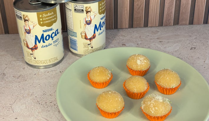 Docinhos claros cobertos com açúcar em forminhas laranja, servidos em prato verde com Leite Moça ao fundo.