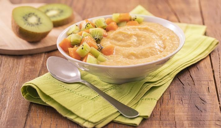 Foto em tons de verde da receita de creminho de laranja servida em um bowl de porcelana branco com uma colher prateada ao lado, sobre um pano verde claro. Ao fundo, há uma tábua de madeira com kiwis cortados em cima