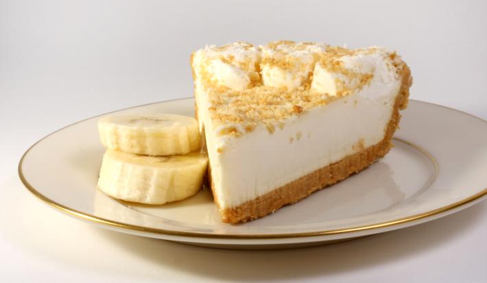 Fotografia de uma fatia de torta de banana e leite MOÇA, com uma farofinha de biscoito e rodelas de banana por cima. A torta está em um prato de vidro branco com as bordas na cor dourado, com duas rodelas de banana ao lado da torta, sobre uma mesa branca.