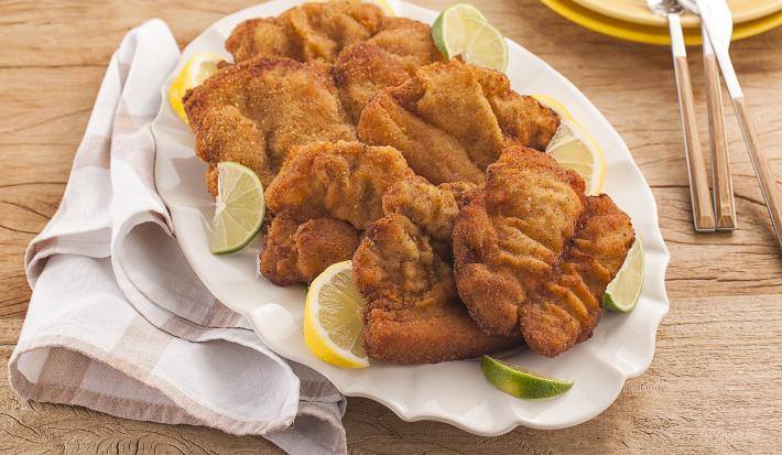 Fotografia em tons de amarelo em uma bancada de madeira clara, um pano branco com listras bege, um prato branco oval com vários bifes suínos à milanesa. Ao fundo, pratos redondos amarelos com talheres em cima deles.