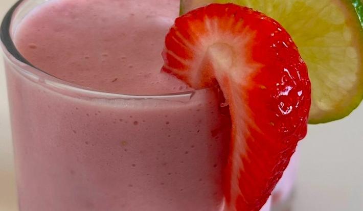 Fotografia de um copo de vidro grande com limonada de morago. Na borda do copo tem uma rodela de limão e um pedaço de morango.