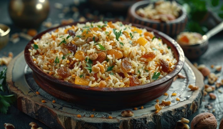 Em cima de uma mesa, tigela com porção de arroz dourado soltinho com uvas passas e nozes picadas.
