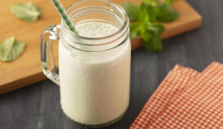 Fotografia em tons de branco, em uma bancada de madeira de cor preta. Ao centro, um copo com o suco de melão com um canudo verde dentro. Ao lado do copo, há um pano quadriculado e ao fundo, uma tábua de madeira de cor marrom folhas de hortelã espalhadas.