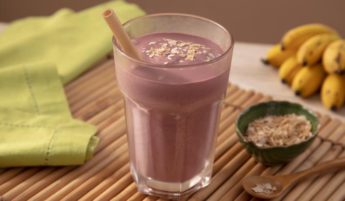 Fotografia em tons de roxo em uma bancada de madeira de cor marrom. Ao centro, um copo contendo o milkshake. Ao lado, há um pano verde, um pote com Neston e ao fundo um cacho de bananas.