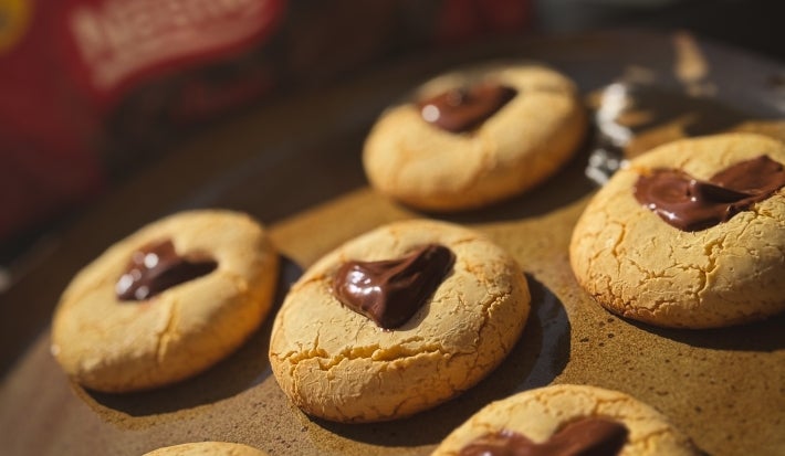 Fotografia de um prato grande e raso de tom marrom na frente da embalagem do chocolate classic meio amargo. Sobre o prato tem alguns sequilhos de leite em pó zero lactose com uma decoração de chocolate em formato de coração.