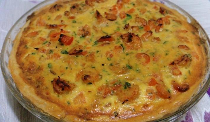 Foto em tons de amarelo da receita de quiche de camarão servida em uma porção grande em uma travessa circular de vidro sobre uma mesa com toalha branca e lilás