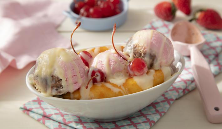 Fotografia em tons de rosa em uma bancada de madeira de cor marrom. Ao centro, uma toalha colorida e em cima, um pote contendo a banana split. Ao lado, uma colher rosa e ao fundo, um pano rosa, morangos e cerejas espalhados.