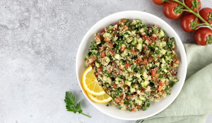 Fotografia em tons claros vista de cima de um recipiente fundo, branco, com uam receita de tabule. O recipiente está ao lado de alguns tomates, um pano verde claro e folhas de salsinha, sobre uma mesa de cimento queimado e tom claro.
