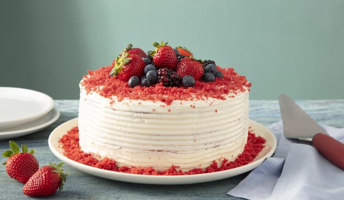 Imagem de um bolo red velvet inteiro decorado com frutas vermelhas. Ao lado, uma espátula, pratos e morangos.