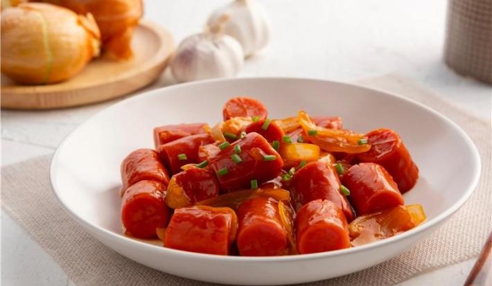 Fotografia em tons claros de um prato de vidro fundo com salsichas em pedaços e um pouco de molho com cebolas cortadas em tiras médias, cozidas no molho também. O prato está sobre um pano fino bege, o qual está apoiado em uma mesa branca.