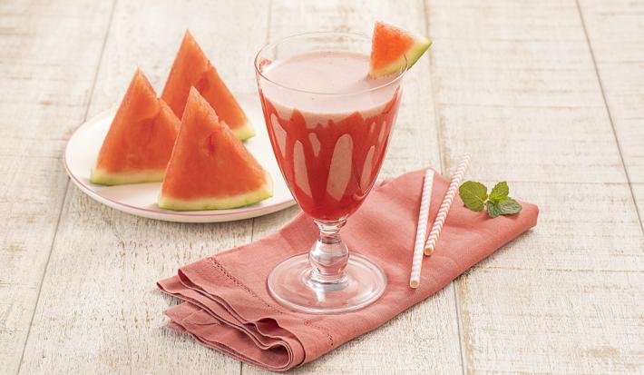 Foto da receita da bebida refrescante de melancia e morango servida em uma taça de vidro com um pedaço de melancia para decorar, há um pano vermelho embaixo da taça e sobre a mesa de madeira clara. Ao fundo um pires branco com três pedaços de melancia