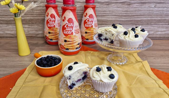 Fotografia em tons de amarelo com dois recipientes de vidro ao centro. Em cima dos recipientes existe vários cupcakes recheados com blueberry e coberto com um creme branco feito de GALAK.