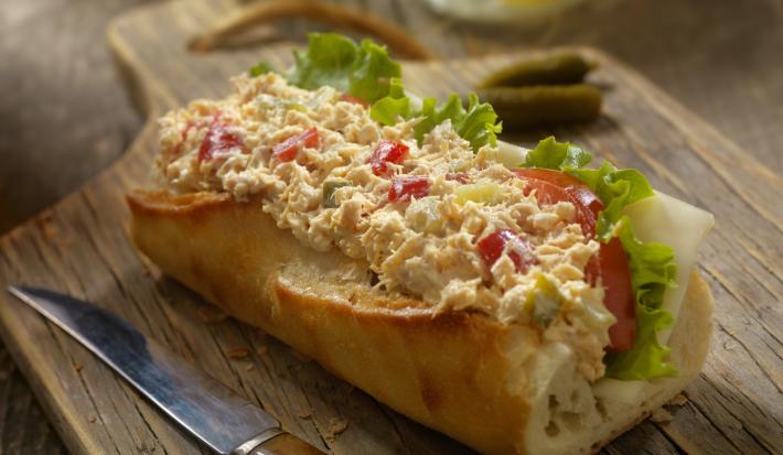 Fotografia de um sanduíche no pão francês com tomate, patê, queijo e alface, sobre uma tábua de madeira escura. Ao lado, uma faca com o cabo marrom.