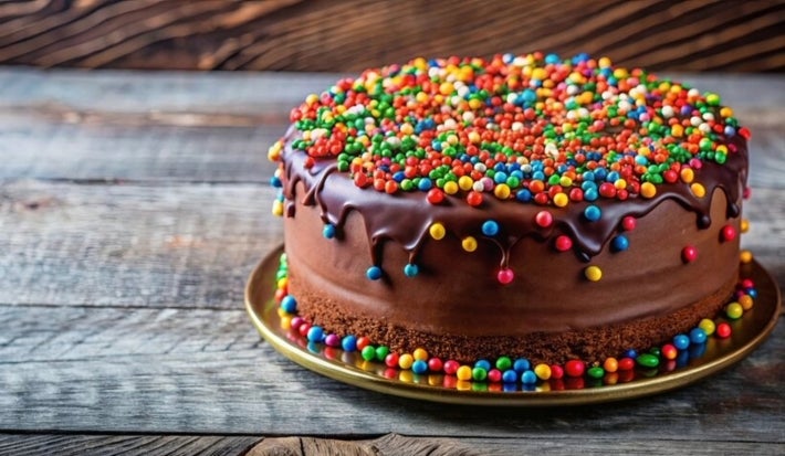 Fotografia de um bolo de chocolate redondo, coberto com uma suave cobertura de chocolate e decorado com coloridos confeitos. O bolo está sobre um prato dourado, contrastando com a superfície de madeira rústica.