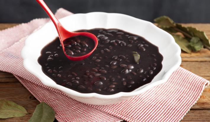 Fotografia em tons de preto e vermelho de uma bancada de madeira, um pano com listras vermelhas e brancas, uma travessa redonda com feijão cozido e seu caldo e uma concha vermelha com um pouco do feijão acima da travessa.