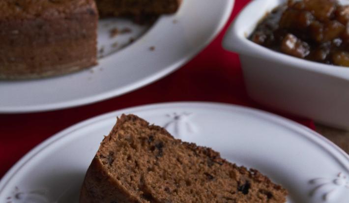 Fotografia em tons de marrom em uma mesa de madeira com um guardanapo vermelho, um prato branco redondo raso com uma fatia de bolo de chocolate com geleia de banana. Ao fundo, um prato maior com o bolo inteiro.