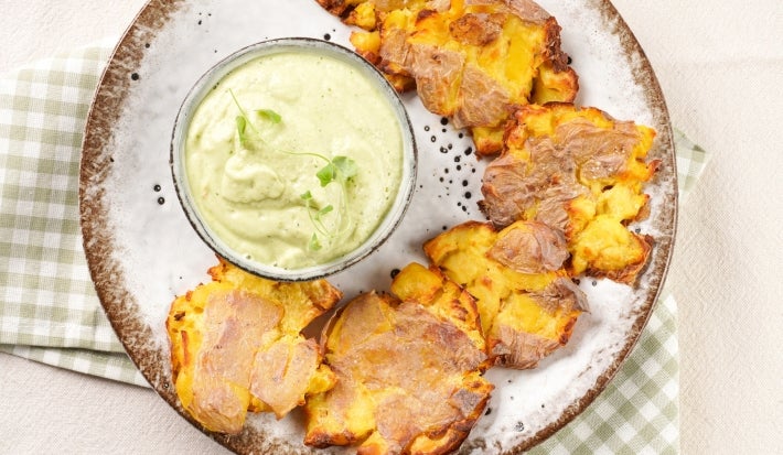 Em cima de uma mesa, prato redondo com batatas ao murro douradas e crocantes e molho verde.