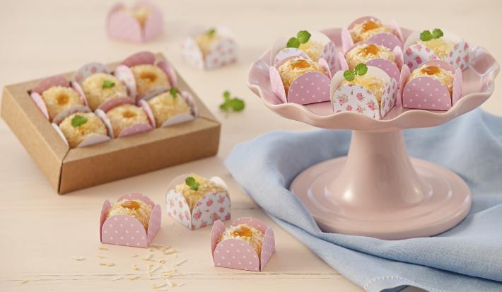 Fotografia em tons de rosa em uma bancada de madeira rosa com um pano azul, um suporte para bolo rosa com alguns brigadeiros brancos com geleia de maracujá em forminhas rosas. Ao fundo, uma caixinha bege com mais brigadeiros brancos.