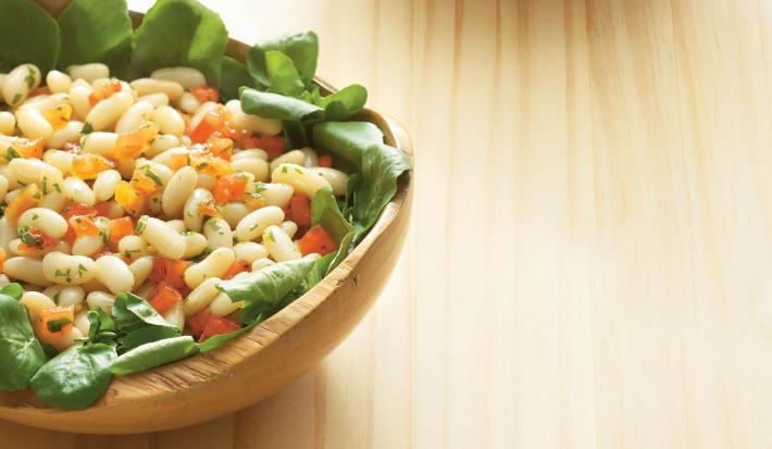 Fotografia de uma mesa de madeira com um bowl de madeira em cima com a salada dentro com folhas, feijão e cenoura.