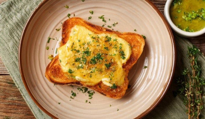 Fotografia em tom claro de uma torrada com creme de queijo parmesão e cebolas, salpicada com salsinha. A torrada está sobre um prato de tom rosa claro.