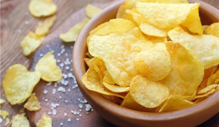 Fotografia de um recipiente médio fundo marrom sobre uma mesa de madeira escura. Dentro do recipiente tem chips de batatas, ao lado tem mais algumas juntamente com um pouco de sal grosso sobre a mesa.