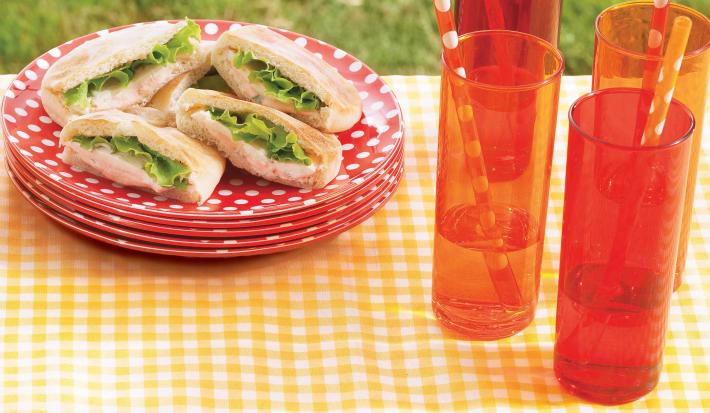 Fotografia em tons de vermelho e amarelo em uma mesa com uma toalha amarela xadrez, pratos vermelhos com bolinhas brancas e os sanduíches de presunto em cima deles. Mais à frente, copos compridos laranjas com suco e canudos de papel.