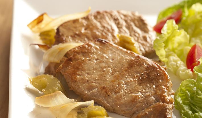 Fotografia em tons de marrom de uma bancada de madeira com um paninho branco e um prato branco com bifes de lombo, pétalas de cebola e salada de alface. Ao fundo uma taça de vidro.