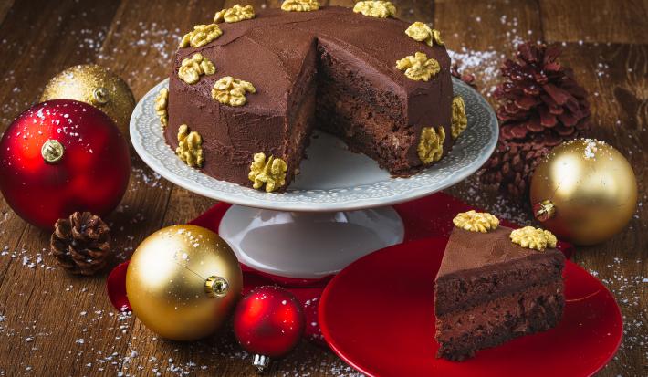 Foto da receita de Bolo Cremoso de Chocolate e Nozes, em um prato branco alto e uma fatia em um prato vermelho. Tudo está em uma bancada de madeira escura decorada com bolas de natal, pinhas e neve artificial