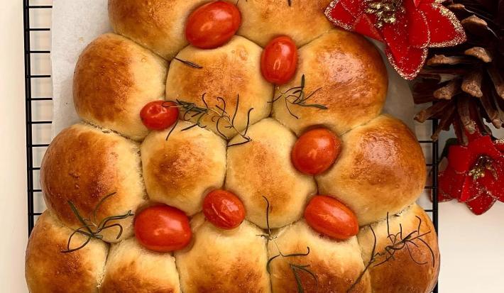 Foto da receita de Pão Caseiro Natalino. Observa-se um pão assado e dourado em formato de árvore, decorado com tomatinhos cereja