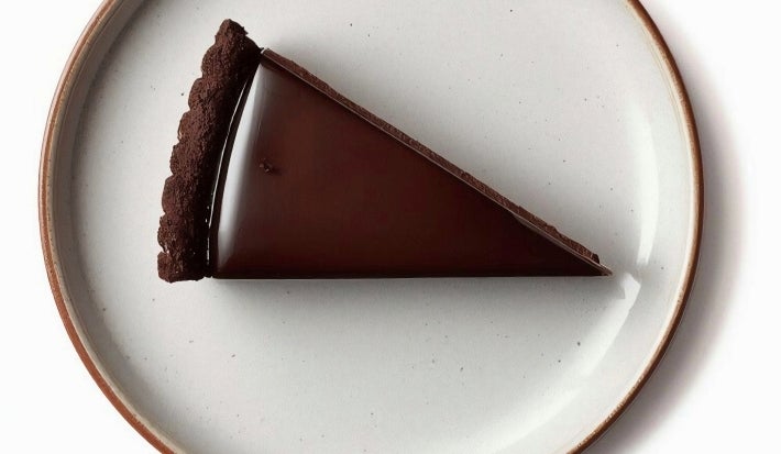 Fotografia vista de cima de uma fatia de torta coberta com chocolate sobre um prato raso claro de vidro.